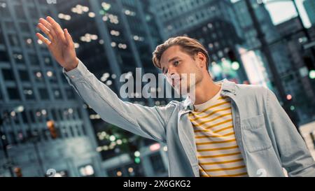 Ritratto di un giovane uomo bello e felice in abiti casual che solleva la mano per prendere un taxi per la strada. Modello maschile di successo nella vita urbana della grande città. Girato con Dutch Angle. Foto Stock