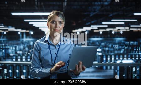 Ritratto di uno specialista IT sorridente che utilizza un computer laptop nel data center, guarda la fotocamera. Ingegnere e-business di successo che lavora in una struttura di cloud computing per aziende agricole di grandi server. Foto Stock