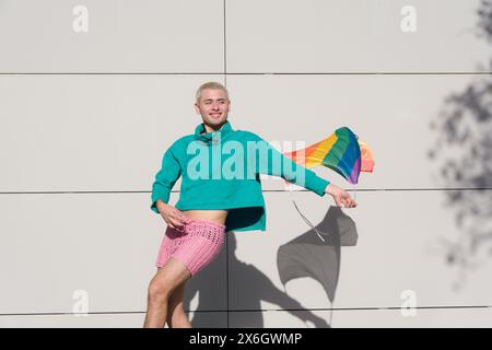 giovane biondo latino con i capelli corti che indossa un maglione verde e pantaloncini rosa che ballano con la bandiera dell'orgoglio all'aperto, è molto orgoglioso di essere gay. Foto Stock