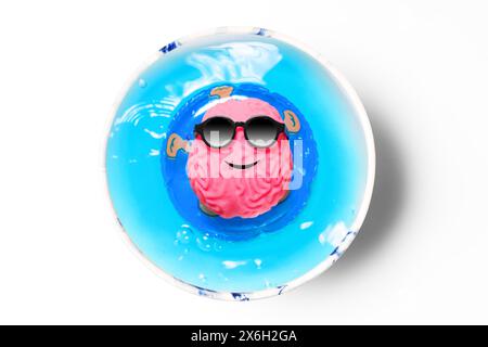 Cervello rosa sorridente adornato da occhiali da sole neri, che galleggia piacevolmente su un anello in una piscina a tazza, su uno sfondo bianco pulito. Felicità creativa, Foto Stock