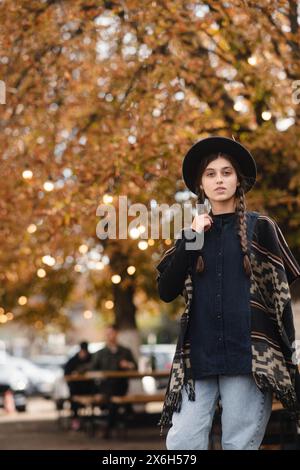 Una ragazza alla moda sfoggia un'eleganza boho, accompagnata da un cappello nero, tra le strade autunnali della città. Foto Stock