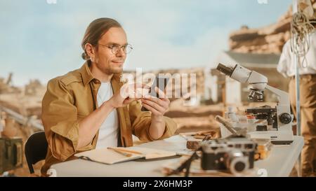 Sito di scavo archeologico: Grande archeologo maschile che fa ricerca, utilizza lo smartphone per condividere la scoperta dei resti fossili, i manufatti della cultura della civiltà antica sui social media di Internet Foto Stock