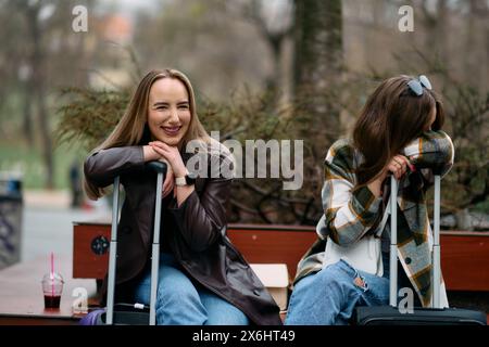 Due donne siedono sulla piazza della città con le valigie, due turisti si siedono e controllano i loro telefoni cellulari sulla piazza della città, un paio di turisti donne Foto Stock