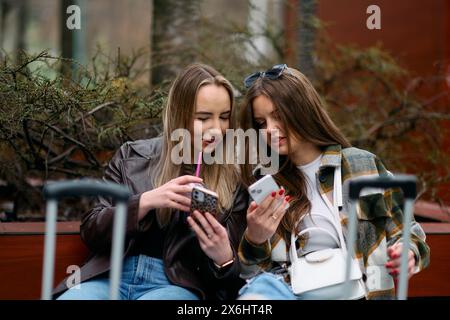 Due donne siedono sulla piazza della città con le valigie, due turisti si siedono e controllano i loro telefoni cellulari sulla piazza della città, un paio di turisti donne Foto Stock