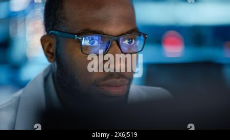 Ritratto del bel Black Man che indossa gli occhiali e lavora con sicurezza su un computer. Giovane ingegnere intelligente maschio o scienziato che lavora in laboratorio. Sfondo blu bokeh. Primo piano Foto Stock