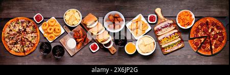 Scena della tavola del cibo spazzatura. Pizza, hamburger, ali di pollo, hot dog e spuntini salati. Vista dall'alto verso il basso su uno sfondo con striscioni in legno scuro. Foto Stock