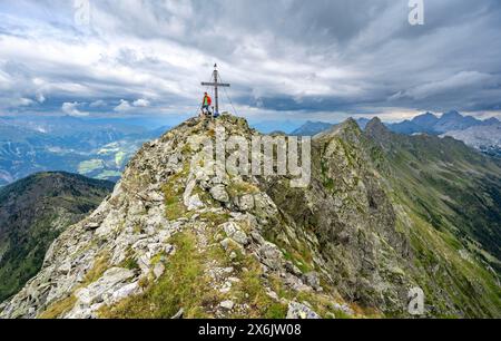 Alpinista sulla cima rocciosa a punta del Raudenspitze o Monte Fleons con croce sommitale, panorama montano sul crinale principale del Carnico, Carnic Foto Stock