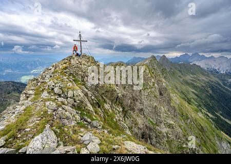 Alpinista sulla cima rocciosa a punta del Raudenspitze o Monte Fleons con croce sommitale, panorama montano sul crinale principale del Carnico, Carnic Foto Stock