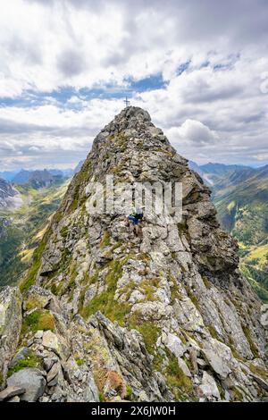 Alpinista su uno stretto sentiero di montagna, cima rocciosa a punta del Raudenspitze o Monte Fleons, con croce a picco, crinale principale Carnic, Alpi Carniche Foto Stock