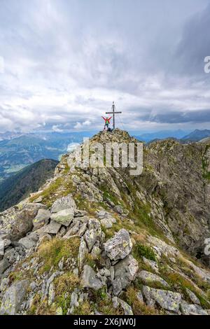 Alpinista stende le braccia in aria, sulla cima rocciosa a punta del Raudenspitze o Monte Fleons con croce sommitale, panorama montano su Foto Stock