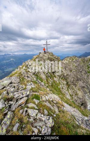 Alpinista sulla cima rocciosa a punta del Raudenspitze o Monte Fleons con croce sommitale, panorama montano sul crinale principale del Carnico, Carnic Foto Stock