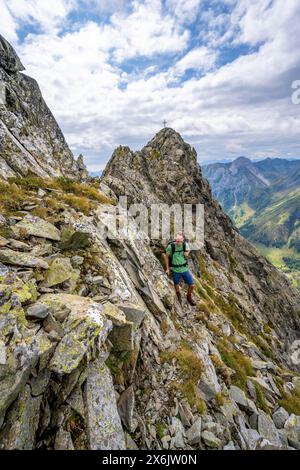 Alpinista su uno stretto sentiero di montagna, cima rocciosa a punta del Raudenspitze o Monte Fleons, con croce a picco, crinale principale Carnic, Alpi Carniche Foto Stock