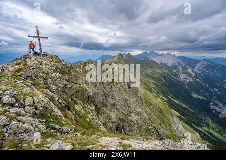 Alpinista sulla cima rocciosa a punta del Raudenspitze o Monte Fleons con croce sommitale, panorama montano sul crinale principale del Carnico, Carnic Foto Stock