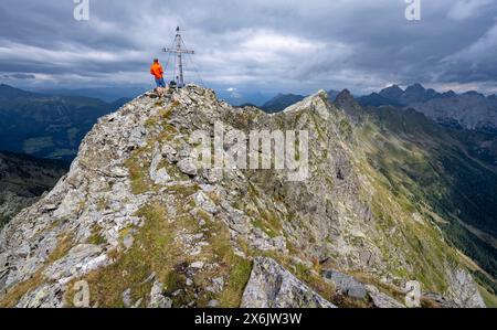 Alpinista sulla cima rocciosa a punta del Raudenspitze o Monte Fleons con croce sommitale, panorama montano sul crinale principale del Carnico, Carnic Foto Stock