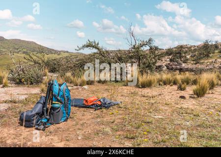 zaino da trekking blu e tenda arancione posizionati sulle rocce di montagna circondati da vegetazione verde e luce naturale e con nuvole di tempesta con le fores Foto Stock