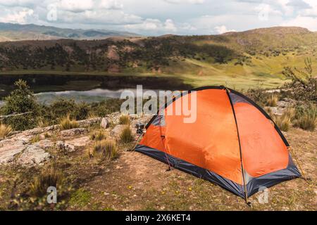 Tenda arancione sulla cima di una montagna con un lago sullo sfondo e cielo blu e nuvole nel mezzo della foresta nella catena montuosa delle Ande Foto Stock