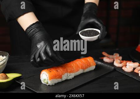 Cuoco in guanti aggiungendo semi di sesamo su gustosi sushi al tavolo di legno nero, primo piano Foto Stock