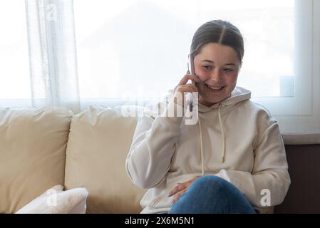Una giovane donna seduta su un divano beige, che si gode una conversazione telefonica in uno spazio luminoso e accogliente Foto Stock