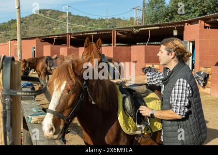 Ritratto di un operaio esperto di stalle adulte in camicia a pipa in piedi vicino al cavallo sellato in un club di campagna Foto Stock