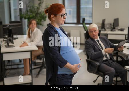 Una donna incinta soffre di dolore e tiene lo stomaco in piedi nel mezzo dell'ufficio accanto ai suoi colleghi. Foto Stock
