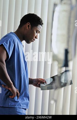 Un chirurgo afroamericano maschio in scrub sta studiando attentamente le informazioni visualizzate su un monitor in una sala operatoria affollata. Il suo espreo focalizzato Foto Stock