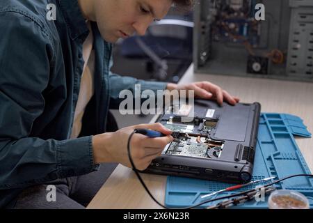 Un tecnico professionista ripristina il notebook utilizzando vari strumenti speciali. Officina di riparazione elettronica, rinnovo tecnologico, affari, riparazioni concettuali. uomo che ripara parti di un computer portatile smontato Foto Stock
