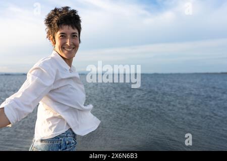 donna dai capelli corti che sorride sulla spiaggia, indossa camicia bianca, top rosso e jeans, cielo blu e mare calmo. Foto Stock