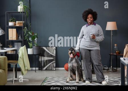 Ritratto a figura intera di donna nera adulta con cane schnauzer in piedi in un interno grigio e che guarda la macchina fotografica pronta per camminare, copia spazio Foto Stock