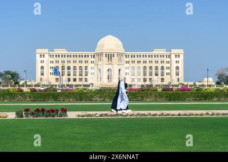 Mascate, Oman - 2 gennaio 2024: Una figura solitaria fa una tranquilla passeggiata nel verde lussureggiante, con la maestosa architettura di un grande edificio Foto Stock