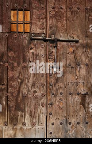 Dettaglio di una vecchia porta in legno ruvido, resistente agli agenti atmosferici, con piccola finestra e serratura a bullone in metallo. Foto Stock