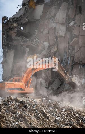 Un escavatore arancione sta attivamente demolendo un grande edificio, creando una nuvola di polvere e detriti nel processo. Foto Stock