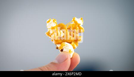 Una persona tiene in mano un gruppo di popcorn ricoperti di caramello, mostrati su uno sfondo blu tenue, che enfatizza la consistenza e il colore dorato dello spuntino. Foto Stock