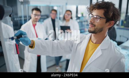 Medical Science Laboratory: Handsome Latin male Scientist scrive analisi dettagliata dei dati di progetto sul Consiglio, il suo team diversificato di colleghi ascolta. Giovani scienziati che risolvono problemi. Foto Stock