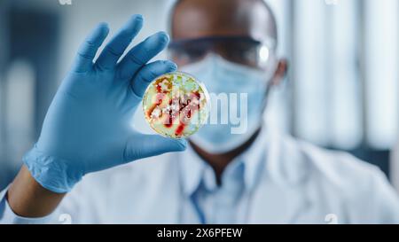 Scienziato maschio afroamericano che indossa maschera e occhiali per il viso che guardano a una piastra di Petri con prodotti chimici geneticamente modificati. Microbiologo che lavora in laboratorio moderno con apparecchiature tecnologiche. Foto Stock