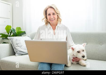 Allegra donna anziana in pensione con un cane e divertirsi con un animale domestico a casa con un portatile Foto Stock