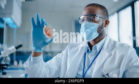 Scienziato maschio afroamericano che indossa maschera e occhiali per il viso che guarda il piatto di Petri con i campioni geneticamente modificati. Microbiologo che lavora in laboratorio moderno con apparecchiature tecnologiche Foto Stock