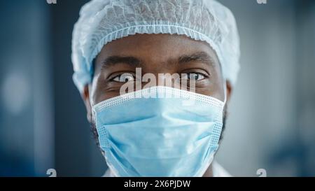 Primo piano Ritratto di un bel medico afroamericano nero o chirurgo che indossa una maschera protettiva e un cappello chirurgico monouso. Scienziato che guarda con calma la fotocamera. Paziente prima dell'intervento chirurgico. Foto Stock