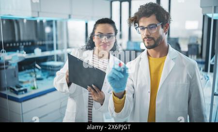 Medical Science Laboratory: Handsome Latin male Scientist scrive Project Data Analysis sul Glass Board, la sua collega Black Female controlla le informazioni sugli Appunti. Giovani scienziati che risolvono problemi Foto Stock