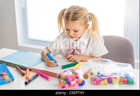 Una piccola studentessa con i capelli intrecciati è seduta alla scrivania con matite colorate Foto Stock