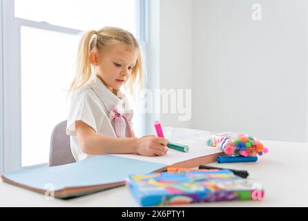 Una piccola studentessa con i capelli intrecciati è seduta alla scrivania con matite colorate Foto Stock