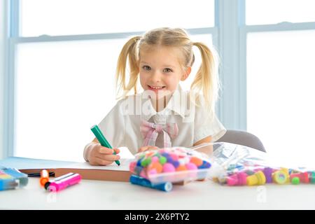 Una piccola studentessa con i capelli intrecciati è seduta alla scrivania con matite colorate Foto Stock