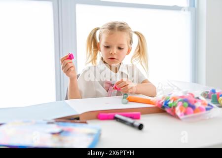 Una piccola studentessa con i capelli intrecciati è seduta alla scrivania con matite colorate Foto Stock