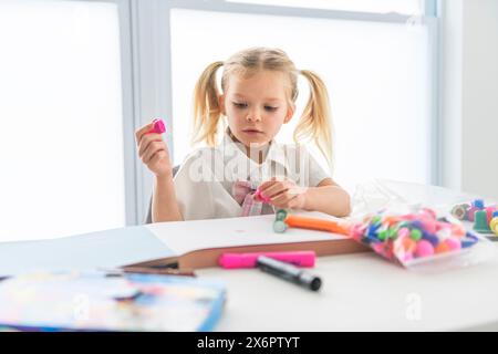 Una piccola studentessa con i capelli intrecciati è seduta alla scrivania con matite colorate Foto Stock