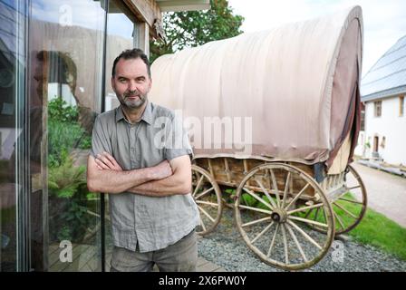 Adorf, Germania. 16 maggio 2024. L'agricoltore biologico Bert Bochmann si trova accanto a un vecchio carro trainato da cavalli pieno di fieno che serve come luogo di riposo per i pellegrini. La fattoria si trova su un sentiero medievale che collega il monastero di Chemnitz, che ora fa parte della via sassone di San Giacomo. L'offerta per gli escursionisti in cerca di illuminazione deve essere ampliata. Tra un anno, le abitazioni portatili per i pellegrini potrebbero trovarsi nei prati di Erzgebirge tra mucche e cavalli. Crediti: Jan Woitas/dpa/Alamy Live News Foto Stock