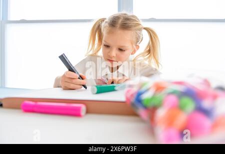 Una piccola studentessa con i capelli intrecciati è seduta alla scrivania con matite colorate Foto Stock