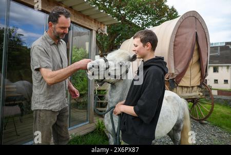 Adorf, Germania. 16 maggio 2024. L'agricoltore biologico Bert Bochmann si trova con il suo pony Eddie e Jane Bochmann accanto a un vecchio carro trainato da cavalli pieno di fieno, che serve come luogo di riposo per i pellegrini. La fattoria si trova su un sentiero medievale che collega il monastero di Chemnitz, che ora fa parte della via sassone di San Giacomo. L'offerta per gli escursionisti in cerca di illuminazione deve essere ampliata. Tra un anno, le abitazioni portatili per i pellegrini potrebbero trovarsi nei prati di Erzgebirge tra mucche e cavalli. Crediti: Jan Woitas/dpa/Alamy Live News Foto Stock