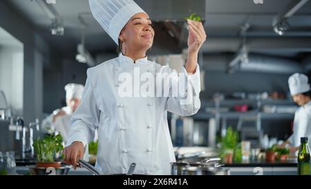 Black Female Chef prende le erbe fresche, le profuma con un sorriso, ingrediente segreto che rende speciale la ricetta della nonna. Cucina tradizionale del ristorante con piatti autentici, cibo salutare e nutriente Foto Stock