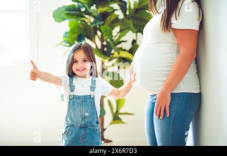 Figlia eccitata che tocca il pancino della mamma incinta, in piedi accanto alla finestra Foto Stock