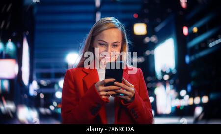 Bella donna in piedi, con smartphone su City Street con luci al neon bokeh che brillano di notte. Bella donna sicura e sorridente che usa il telefono cellulare. Ritratto cinematografico con tracciamento medio. Foto Stock