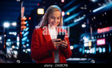 Bella donna in piedi, con smartphone su City Street con luci al neon bokeh che brillano di notte. Bella donna sicura e sorridente che usa il telefono cellulare. Ritratto cinematografico con tracciamento medio. Foto Stock